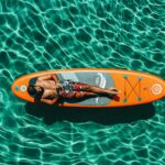 Relaxed man sunbathing on colorful paddleboard in clear blue water.