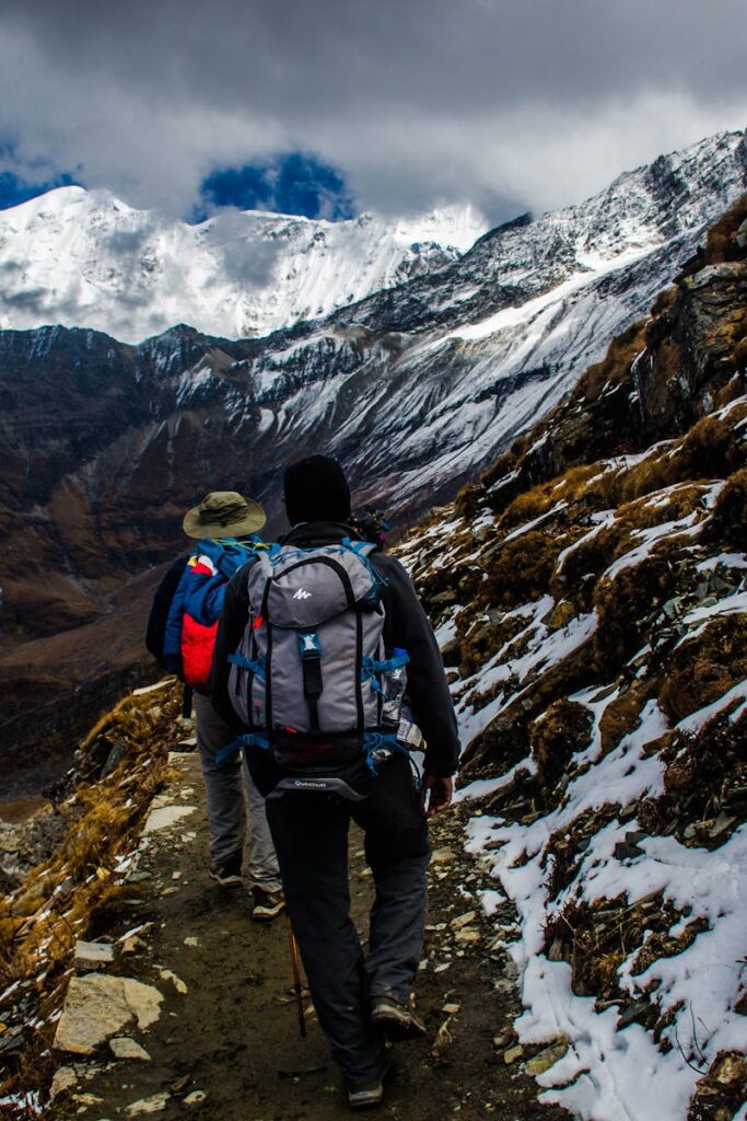 Two hikers navigating a snowy trail in rugged mountainous terrain under dramatic skies.